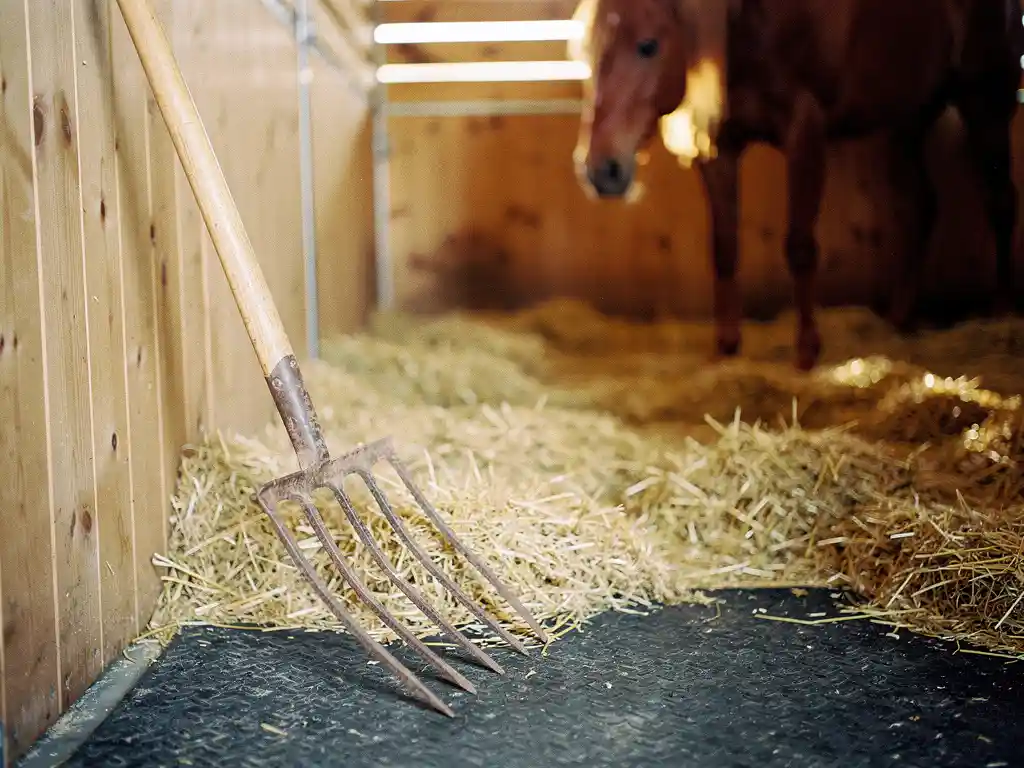Weathered wooden pitchfork leaning against a horse stall wall, fresh golden straw covering a dark rubber mat, chestnut horse softly blurred in background.
