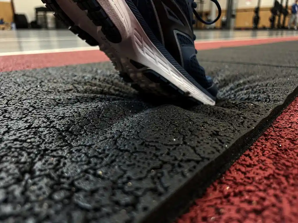 Athletic shoe pressing into a textured rubber sports mat mid-stride, with a blurred gymnasium floor in the background.